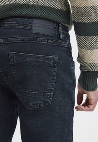 Tamno plavi trapkasti traperici s dodatnom zakovicom, s blagom teksturom i laganim izblješnjenjem. Oznaka brenda vidljiva na pojasu.