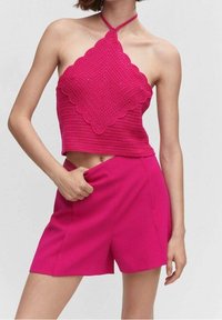 Kobieta w gorącym różowym, szydełkowym halter crop topie z falbaniastymi brzegami oraz dopasowanych szortach z wysokim stanem, pozująca z ręką w kieszeni.