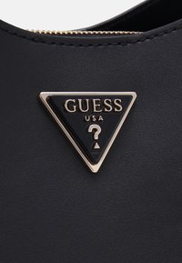Matériau texturé noir présentant un logo Guess triangulaire avec un accent doré sous la forme d'un point d'interrogation, sur un fond sombre.
