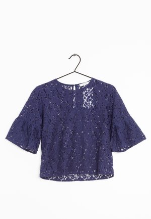 Marineblå blondebluse med blomsterprint, halværmer, rund halsudskæring og løstsiddende pasform hængende på en sort bøjle.