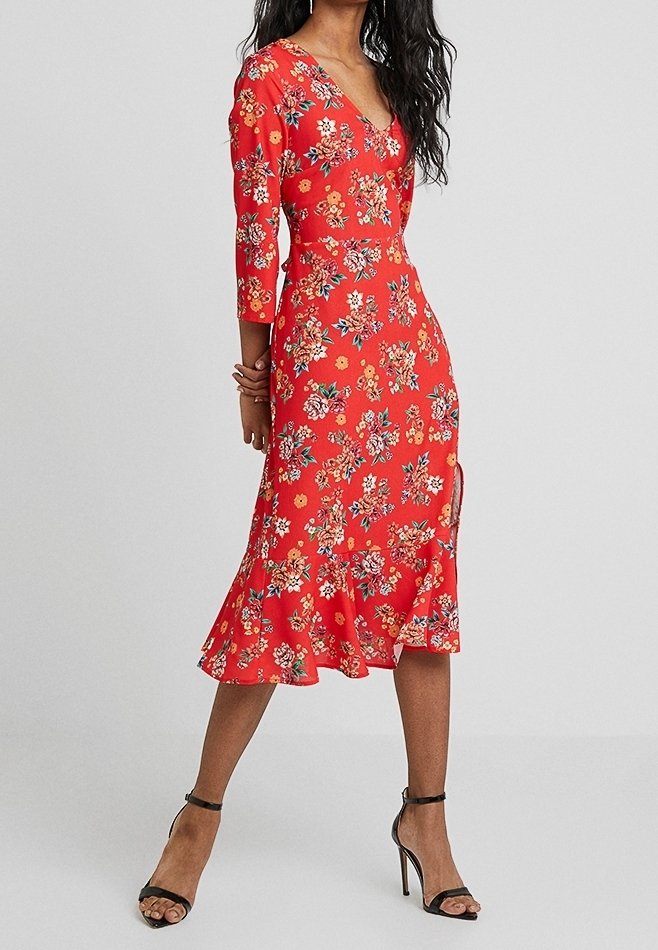 Femme portant une robe midi rouge à fleurs avec des manches trois-quarts et un décolleté en V, associée à des sandales à talons aiguilles noires avec bride à la cheville.