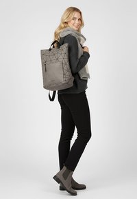 Pilkas geometrinio rašto kuprinė su viršutine rankena, šoniniu užtrauktuku ir reguliuojamomis petnešomis. Nešiojama ant juodos megztinio ir kelnių, su pilkomis aulinėmis bateliais.