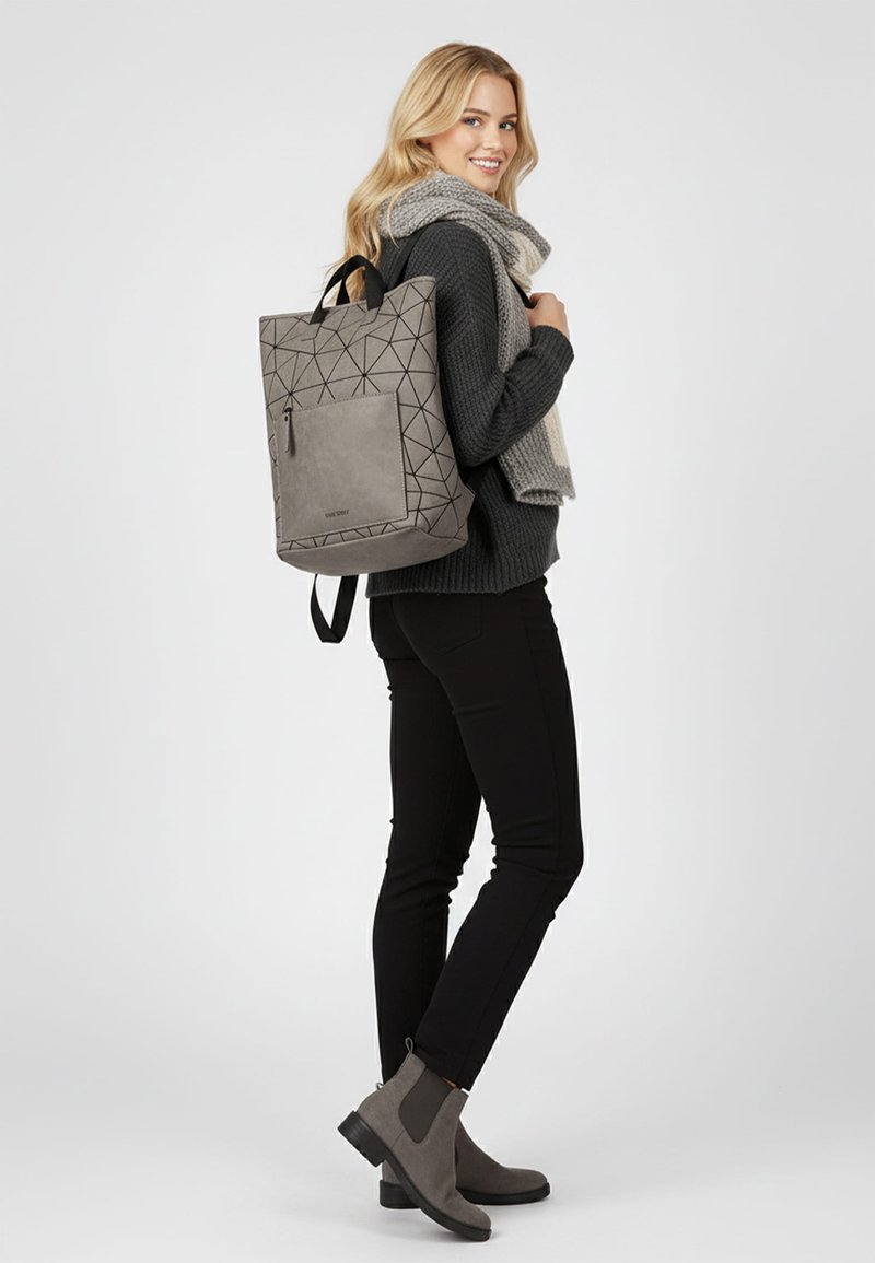 Pilkas geometrinio rašto kuprinė su viršutine rankena, šoniniu užtrauktuku ir reguliuojamomis petnešomis. Nešiojama ant juodos megztinio ir kelnių, su pilkomis aulinėmis bateliais.