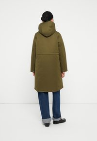 Parka verde oliva con cappuccio, caratterizzata da una vestibilità dritta, maniche lunghe e dettagli con bottoni. Indossata sopra jeans blu risvoltati.