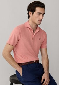 Hombre con una camisa polo color coral con un pequeño logo, pantalones azul marino y un cinturón tejido marrón, sentado en un taburete negro contra un fondo liso.