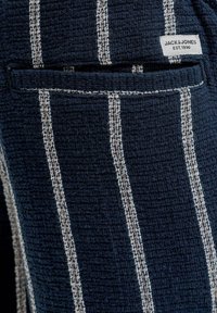 Tissu bleu foncé texturé avec des rayures blanches verticales et une petite étiquette "JACK & JONES EST. 1990" près d'une poche à rabat.
