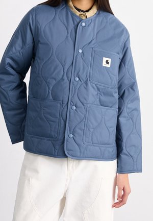 Persoon draagt een blauwe doorgestikte jas met drukknopen en drie voorzakken, gecombineerd met een witte broek.