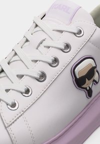 Λευκό αθλητικό παπούτσι με λευκά κορδόνια, μεταλλικά περβάζια, μοβ σόλα και έμβλημα καρτούν με φιγούρα που φορά γυαλιά ηλίου στο πλάι.