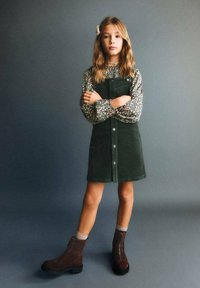 Zelené kordové šaty s předními knoflíky, doplněné květinovou blůzou s dlouhým rukávem. Hnědé kotníkové boty a šedé pozadí dotvářejí vzhled.