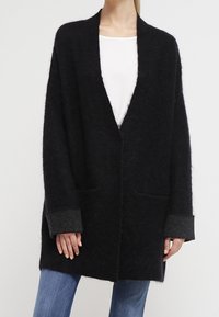Cardigan oversize nero realizzato in un materiale morbido e texture. Presenta una scollatura aperta, due tasche e polsini grigi.