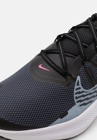 Schwarzer Sportschuh mit einer strukturierten Mesh-Oberfläche, die ein pinkfarbenes Nike-Logo zeigt, komplexen Schnürungen und einer weißen Außensohle mit einem weichen, gerippten Design.