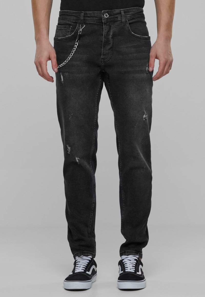 Jeans de mezclilla negra con corte slim, que presentan detalles desgastados y un acento de cadena desmontable en el lazo del cinturón. Llevados con zapatillas negras.