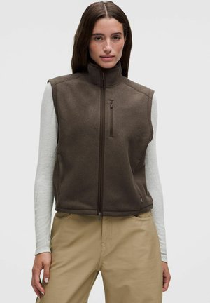 Frau mit langen dunklen Haaren, die eine braune Zip-Up-Weste über einem hellgrauen Langarmshirt und beigen Hosen trägt, steht vor einem einfachen Hintergrund.