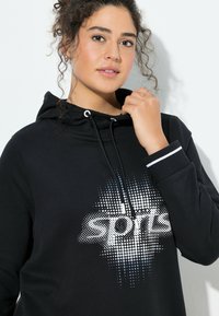 Felpa nera con cappuccio e coulisse, con stampa bianca e puntini blu che formano la parola "sports". I polsini hanno una striscia accentata bianca.