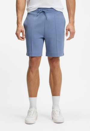 Uomo con pantaloncini azzurri con coulisse, scarpe da ginnastica bianche e calzini bianchi alti, in piedi su sfondo bianco semplice.