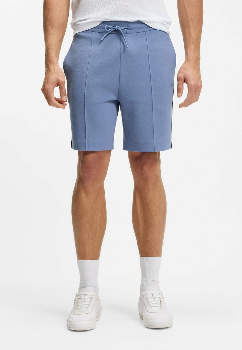Man draagt lichtblauwe shorts met trekkoord, witte sneakers en witte enkelsokken, staand tegen een effen witte achtergrond.