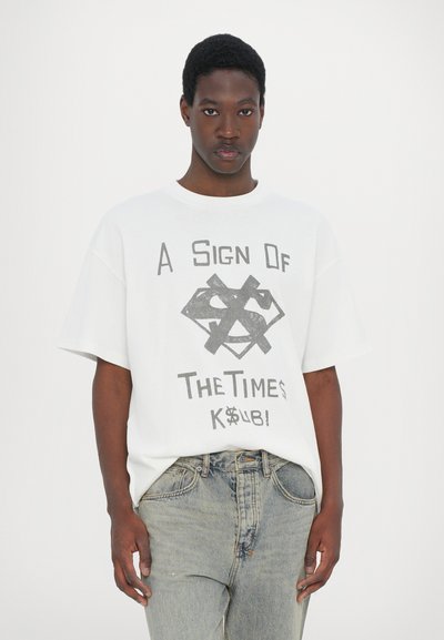 White cotton t-shirt featuring a graphic design with text and symbols in dark gray. Relaxed fit with dropped shoulders, paired with light denim jeans.