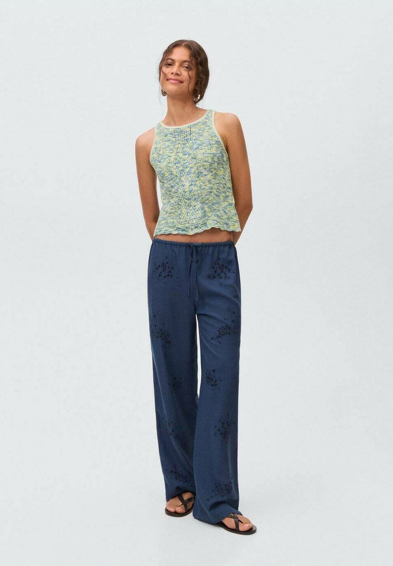 Green and yellow patterned knit crop top with scalloped hem paired with loose navy pants featuring floral embroidery, worn with black flip-flops.