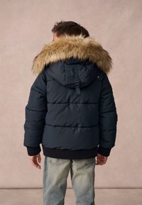 Chaqueta acolchada azul marino con capucha de piel sintética desmontable, puños acanalados y dobladillos elásticos, llevada sobre jeans de tonos azul claro, vista desde la parte de atrás.