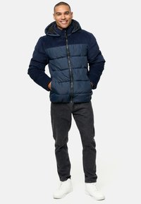 Navy puffer jacket with horizontal stitching, combined with a solid navy top section. Features a hood and zip closure. Worn with dark jeans and white sneakers.