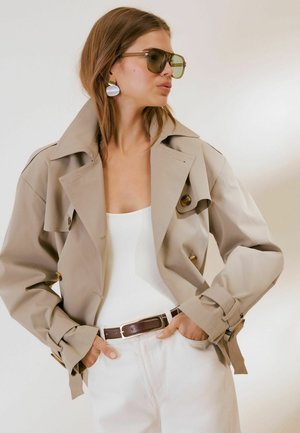 Kvinde iført beige trenchcoat, hvid top, hvide bukser, mørkt bælte, store øreringe og solbriller, poserer med hænderne i lommerne.