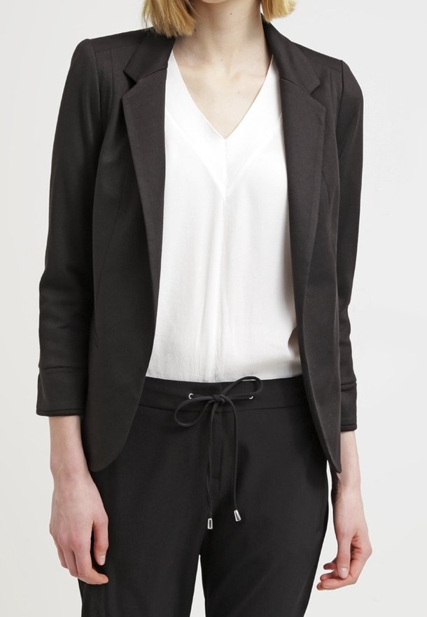 Personne portant un blazer noir cintré par-dessus un chemisier blanc à col en V et un pantalon noir avec cordon, debout devant un fond clair uni.