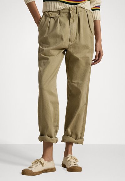 Isik, kes kannab ülespööratud beeže pükse, kreemikaid tenniseid pruunide talladega ja kreemikat kampsunit värviliste triipudega varrukate otsas.