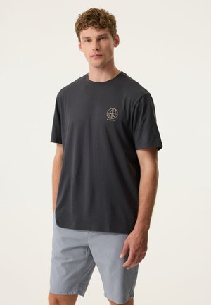 Jonge man met krullend haar die een zwart T-shirt met korte mouwen en logo en lichtgrijze korte broek draagt, staand tegen een effen achtergrond.