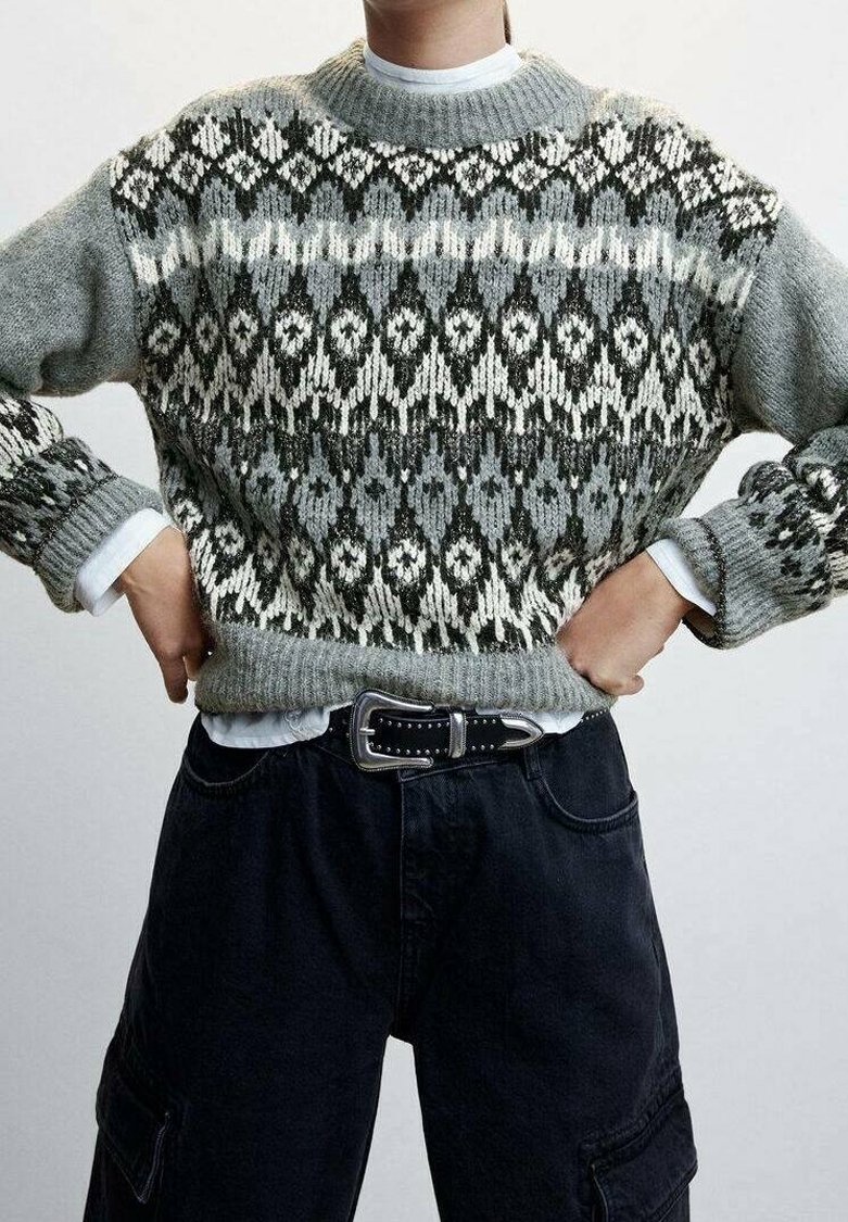 Personne portant un pull en maille gris à motifs sur une chemise blanche, associé à un pantalon cargo noir et une ceinture cloutée noire.
