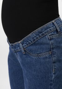 Raskaana olevan vatsa mustassa paidassa näkyvissä löysän sinisen denimin äitiysfarkkujen vyötärön yläpuolella.