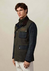 Mladic u zeleno-tamno plavom sakou s džepovima preko tamnozelene rolke, stoji uz jednostavnu bež pozadinu.