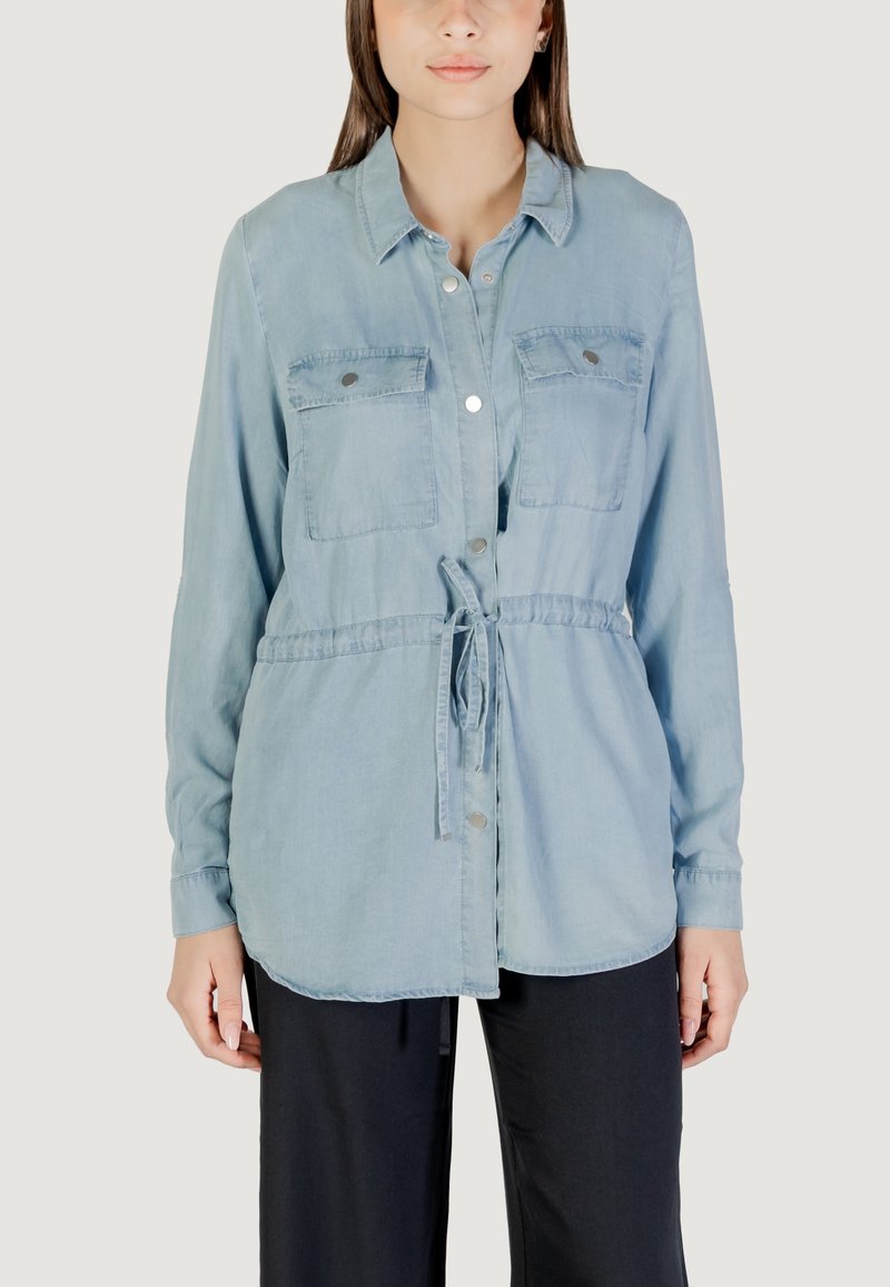 Camisa de mezclilla azul claro con dos bolsillos en el pecho, cierre de botón, cinta ajustable en la cintura y mangas largas. Textura suave, corte relajado.