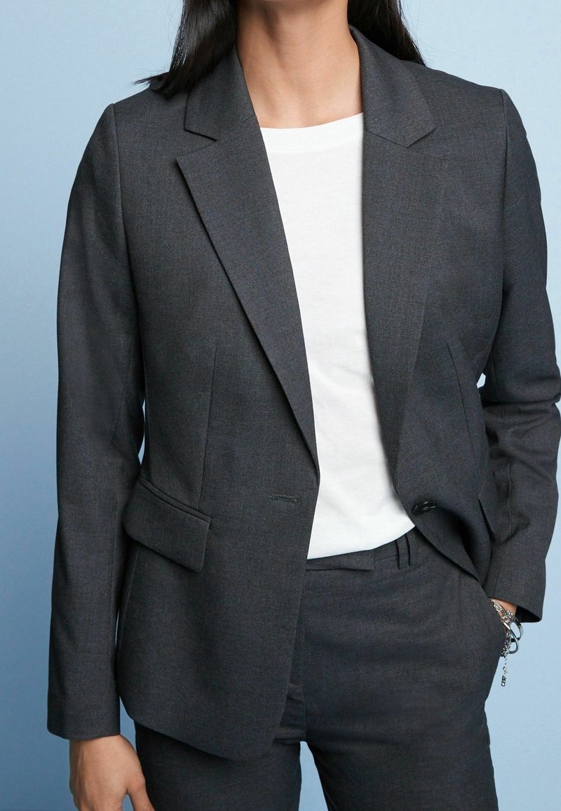 Blazer gris foncé sur mesure avec une texture lisse, fermeture à un bouton et deux poches avant, porté avec une chemise blanche en dessous.