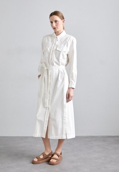 Balta, ilgomis rankovėmis suknelė marškiniai su sagčių užsegimu, dviem krūtinės kišenėmis ir diržu. Dėvima su rudos platformos šlepetėmis ant pilko grindinio.