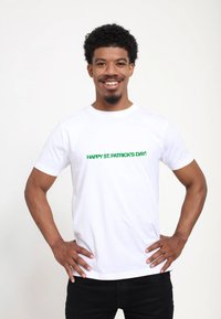 White short-sleeve t-shirt featuring green text that reads "HAPPY ST. PATRICK'S DAY!" displayed on a smiling model with hands on hips.