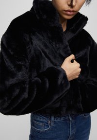 Person iført en sort plysch faux fur jakke, som holder kraven med den ene hånd, kombineret med blå denim jeans.