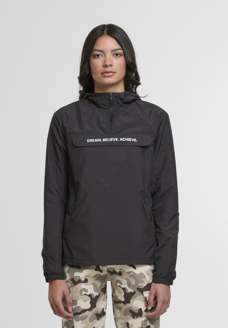 Donna che indossa una giacca nera con zip con la scritta "DREAM. BELIEVE. ACHIEVE." e pantaloni cargo beigi con camouflage, in piedi davanti a uno sfondo semplice.