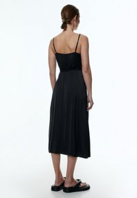 Schwarzes Midi-Kleid mit dünnen Trägern, tailliertem Oberteil und fließendem Rock, aus glattem Stoff gefertigt. Trägt flache schwarze Sandalen. Rückansicht gezeigt.