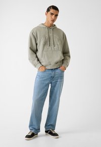 Svetlo zelená mikina s kapucňou a prednou kapsou, vyšitou logom, v kombinácii so svetlomodrými voľnými džínsy a čiernymi teniskami s bielymi akcentmi.