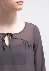 Genomskinlig grå blus med rund halsringning, detaljer med knytning fram och upptagen tyg kring axlarna. Långa ärmar med en mjuk textur.