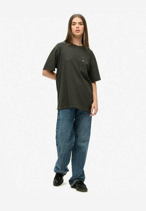 Przesadny ciemnoszary t-shirt z kieszonką, w zestawieniu z niebieskimi dżinsami. Buty są czarne o błyszczącej powierzchni. Gładkie białe tło.