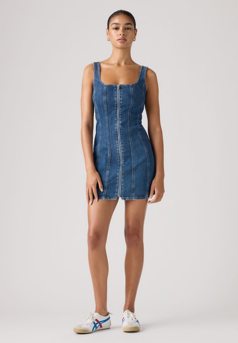 Femme debout portant une mini robe en denim sans manches avec fermeture éclair à l'avant et des baskets blanches avec des rayures bleues et rouges sur un fond clair.