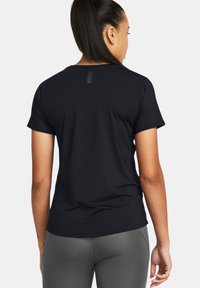 Mujer con cabello largo y oscuro en una coleta, que lleva una camiseta negra perforada de manga corta y Leggings grises, vista desde atrás.