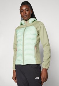 Casaco puffer verde-claro e oliva com fecho de correr, capuz e detalhes em rosa. Apresenta um painel frontal texturizado e mangas ajustadas.