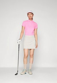 Kobieta w różowej koszulce polo, beżowej spódnicy i daszku do golfa, trzymająca kij do golfa, stojąca na jasnym, jednolitym tle.