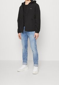 Schwarze Zip-Jacke mit Kapuze, aus glattem Material. Getragen über einem schwarzen Shirt, kombiniert mit hellblauen Jeans und weißen Sneakers.
