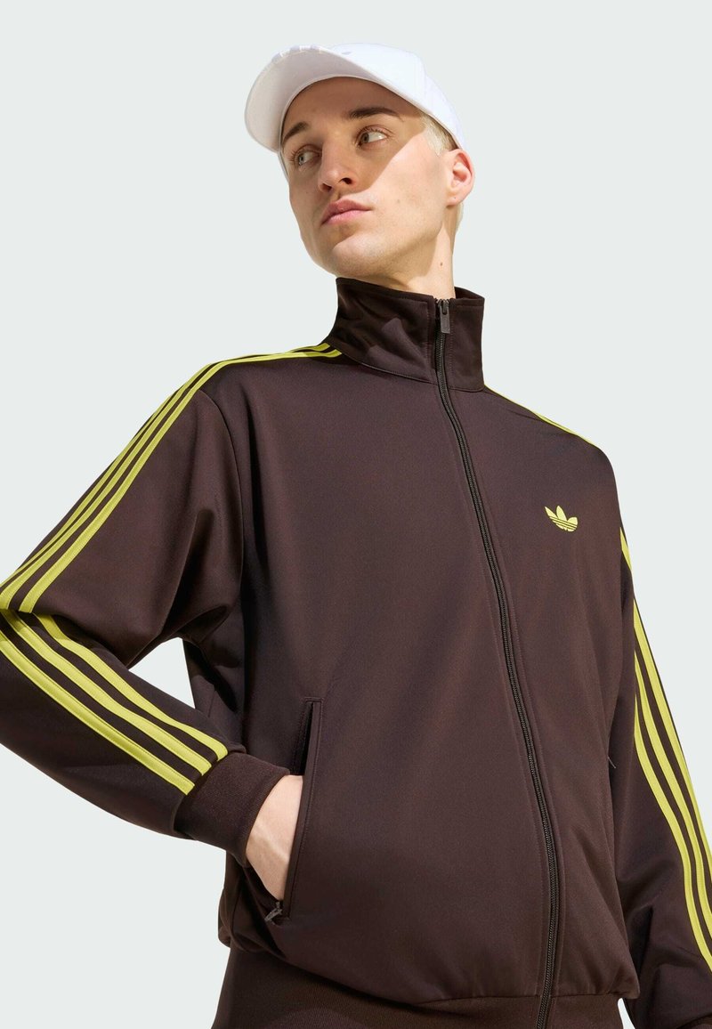 Giovane uomo che indossa una giacca nera con zip con strisce gialle e logo, e un cappello bianco, che guarda di lato con la mano in tasca.