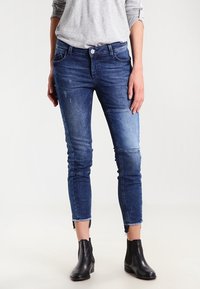 Úzké džíny v tmavě modré barvě s vybledlým designem, s mírným opotřebením a surovým lemem. Spárované s černými kotníkovými botami.