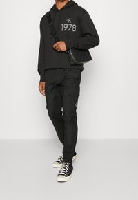Svart hoodie med "CK 1978"-logotyp, tillsammans med svarta cargo-byxor med fickor och svarta sneakers med vita snören. Svart axelremsväska.