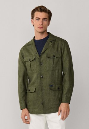 Junger Mann trägt eine olivgrüne, geknöpfte Jacke mit vier Klappentaschen über einem dunkelblauen Hemd und weißen Hosen, steht vor schlichtem Hintergrund.